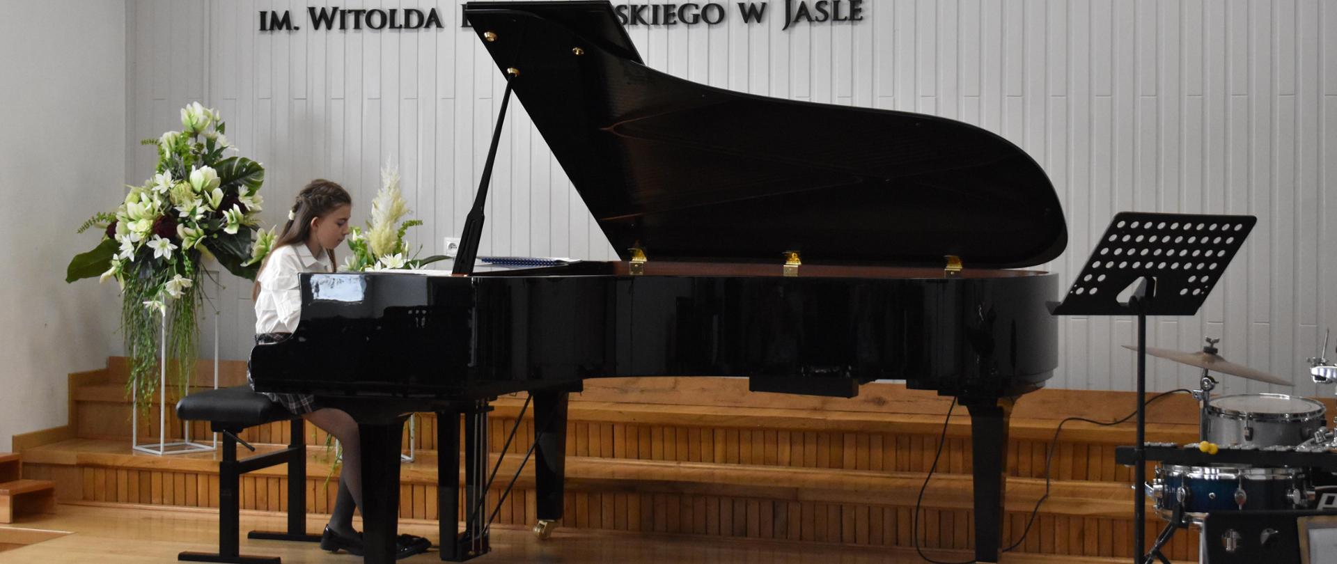 na zdjęciu, w auli PSM, podczas koncertu w trakcie Dni Otwartych PSM I st. w Jaśle widnieje uczennica grająca na fortepianie. Kolorystyka zdjęcia jest biało-brązowo-zielono-czerwono-czarna.