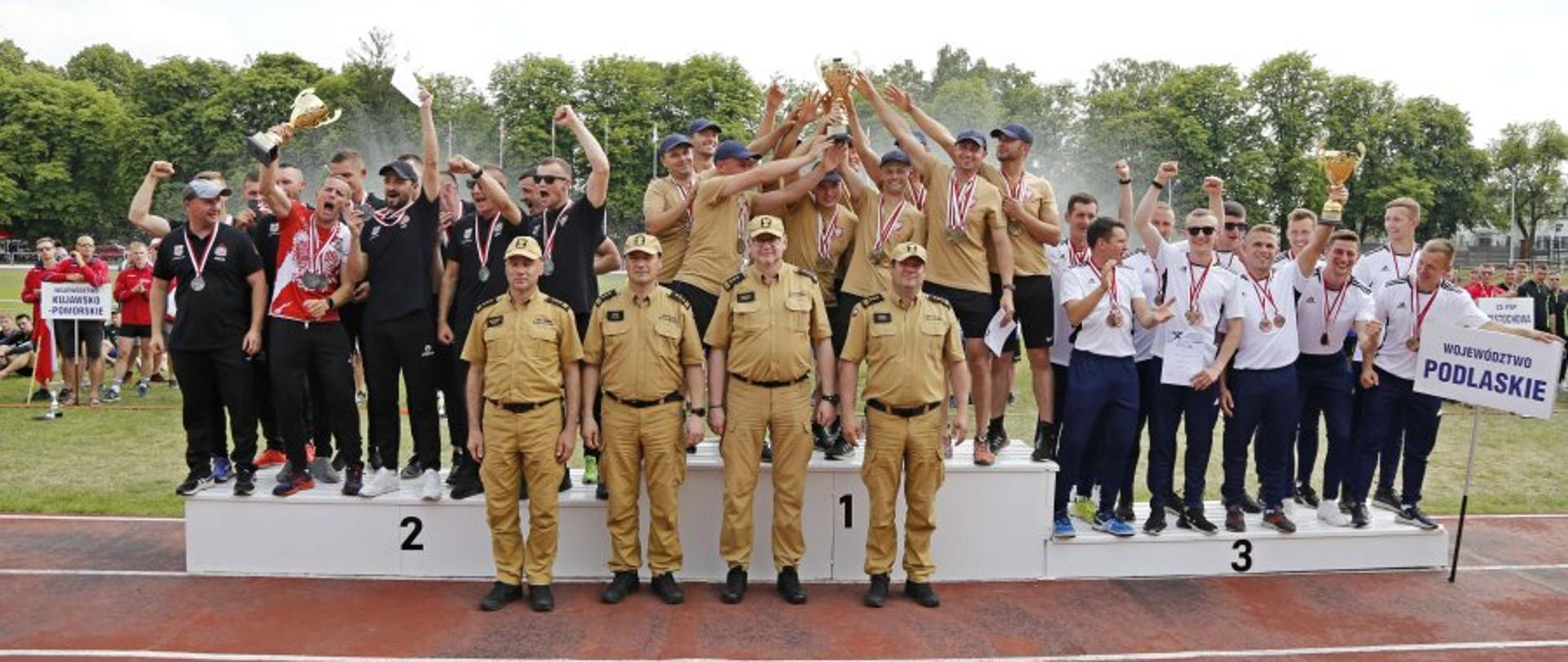 Ceremonia wręczenia medali. Na podium widzimy trzy drużyny wojewódzkie celebrujące otrzymanie medali. Przed podium stoi czterech strażaków w piaskowych mundurach. 