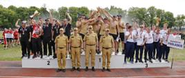 Ceremonia wręczenia medali. Na podium widzimy trzy drużyny wojewódzkie celebrujące otrzymanie medali. Przed podium stoi czterech strażaków w piaskowych mundurach. 