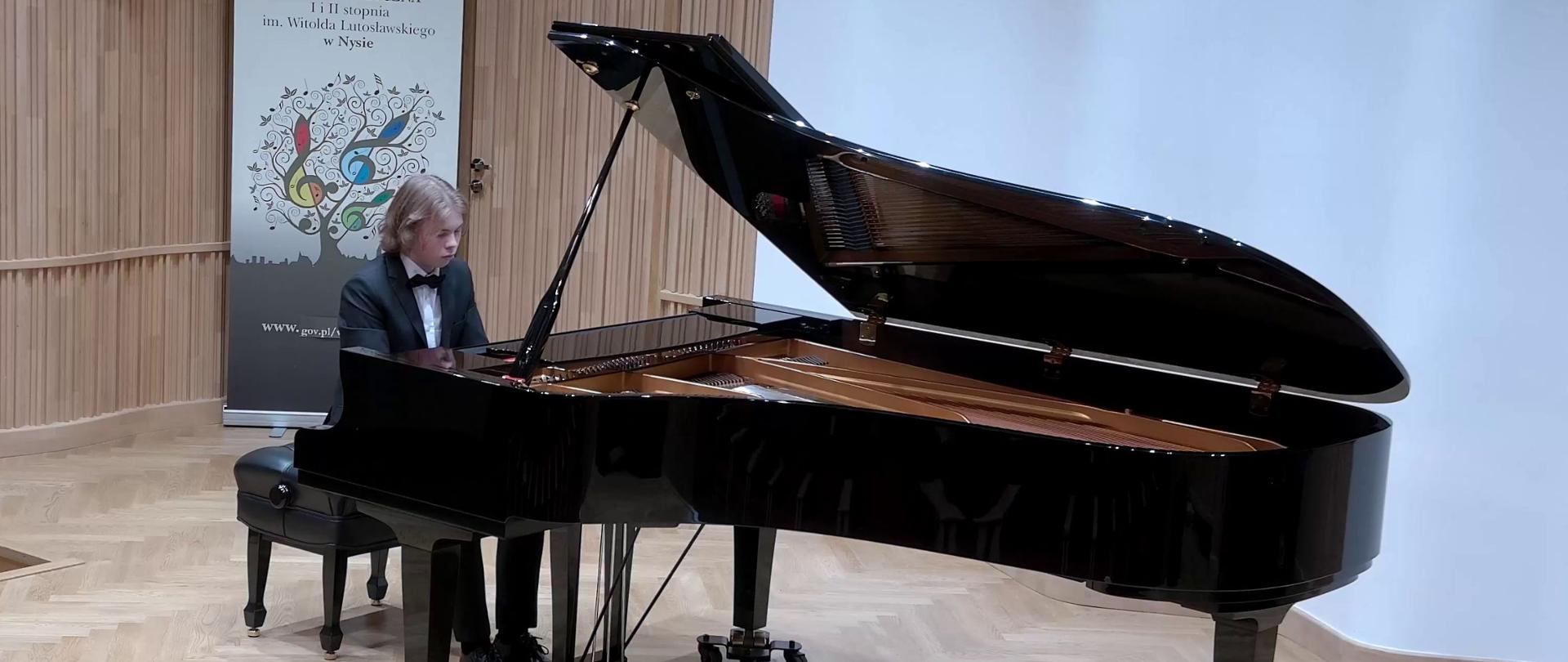 Zdjęcie. Na estradzie sali koncertowej pianista gra na dużym czarnym fortepianie. W tle baner PSM Nysa.