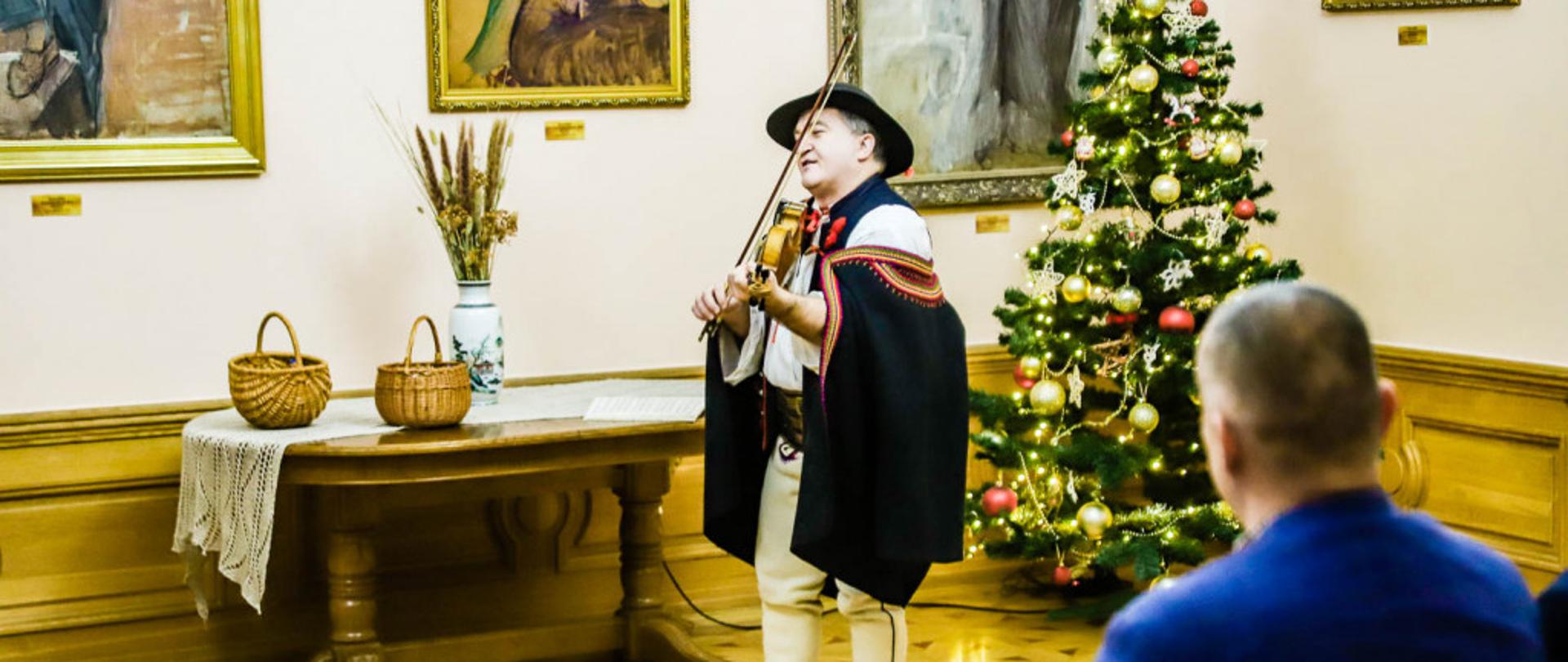 zdjęcie przedstawia Marcina Pokusę w stroju góralskim, w jednej z sal Muzeum Regionalnego w Mielcu, pomalowanej na biało z drewnianą podłógą, w tle widać fragmenty powieszonych na ścianach obrazów oraz ubraną choinkę