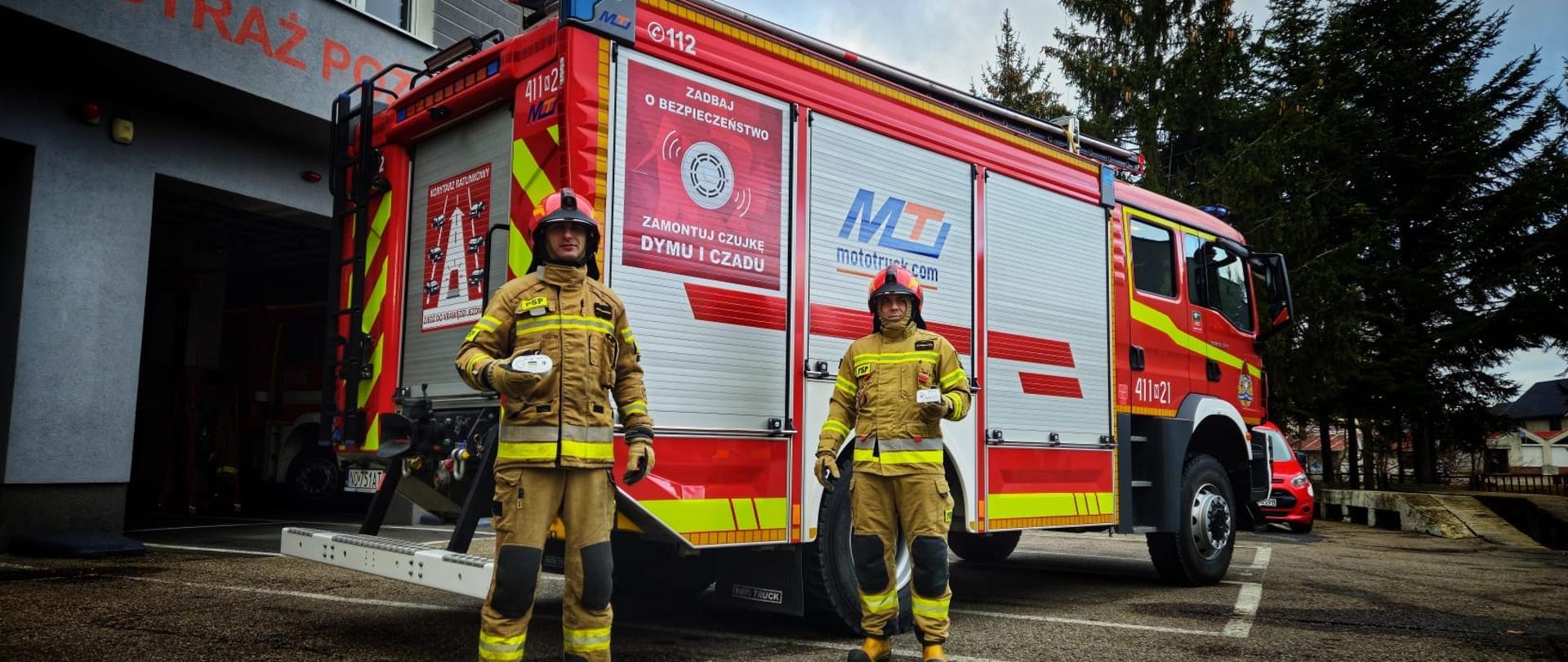 Dwóch strażaków w czerwonych hełmach przy dużym czerwonym czerwonym samochodzie strażackim.