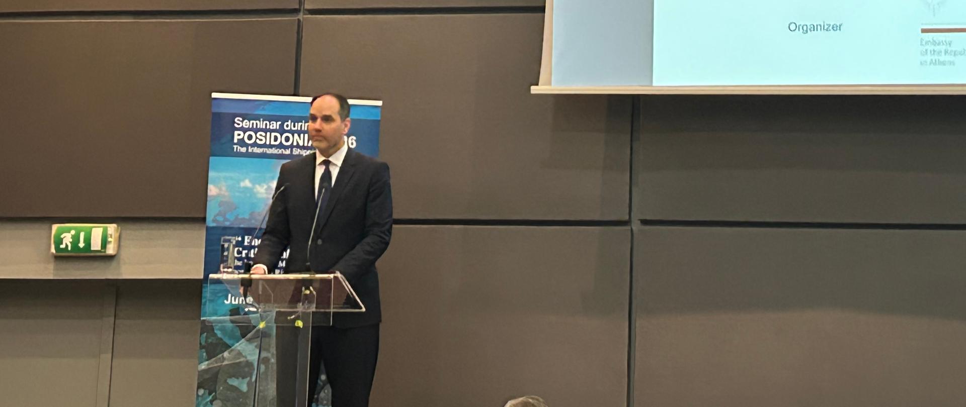 Secretary of State Ignacy Niemczycki speaks at a podium during the “POSIDONIA 2026”. Behind him, a large screen displays the event agenda and keynote speakers, while conference attendees are seated in the audience.