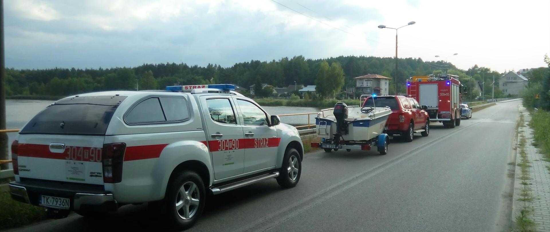 Zdjęcie przedstawia samochody służb ratowniczych stojących na pasie jezdni wzdłuż brzegu zalewu. 