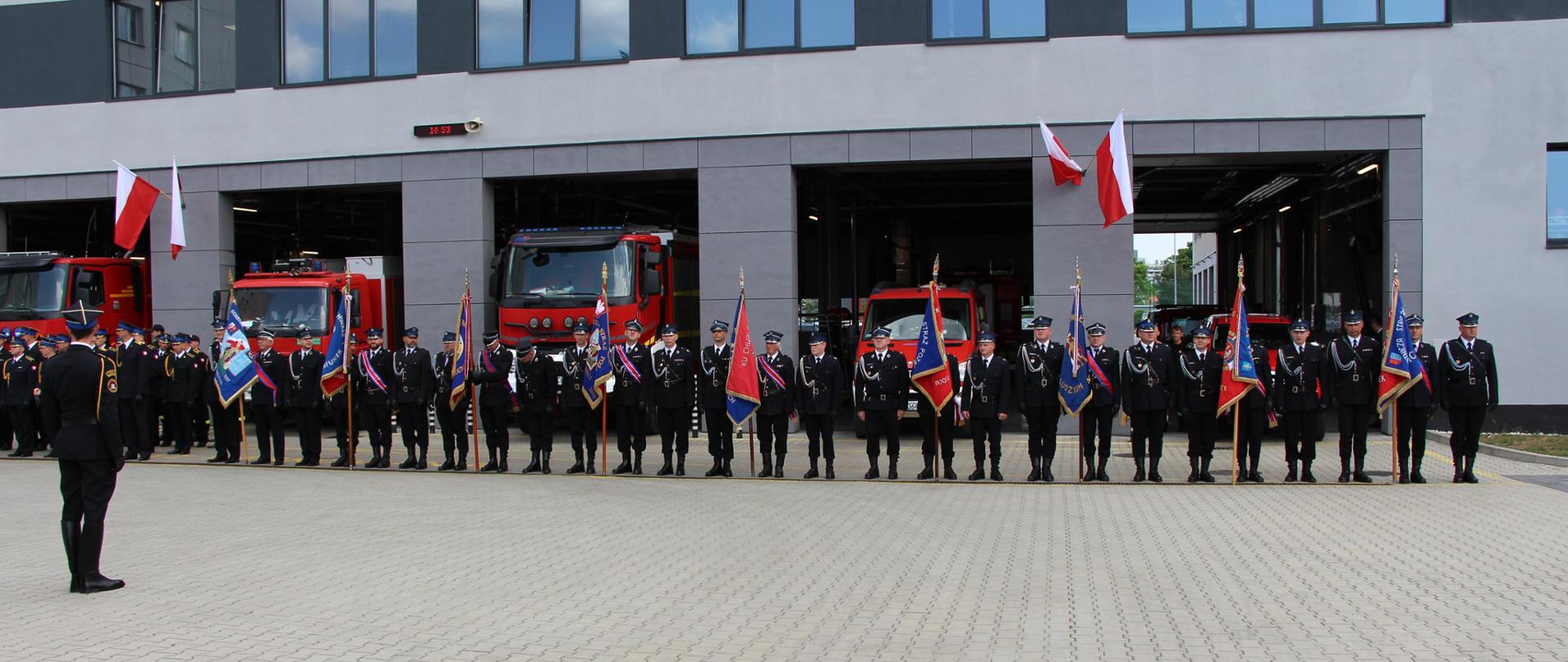 Przed budynkiem Jednostki Ratowniczo-Gaśniczej Numer 1 w Kielcach z otwartymi bramami, na tle zaparkowanych wewnątrz samochodów stoją poczty sztandarowe.
