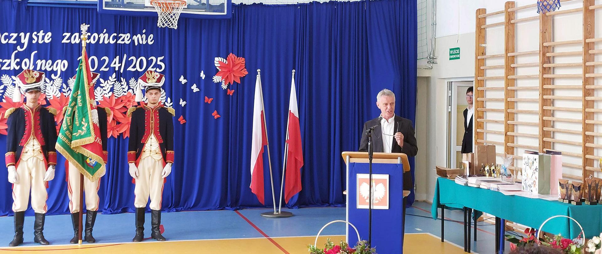 Od lewej: stoi trzech mężczyzn w mundurach, jeden trzyma sztandar, jeden mężczyzna w garniturze przemawia z mównicy, po prawej stoliki przykryte zielonym suknem, na których stoją upominki