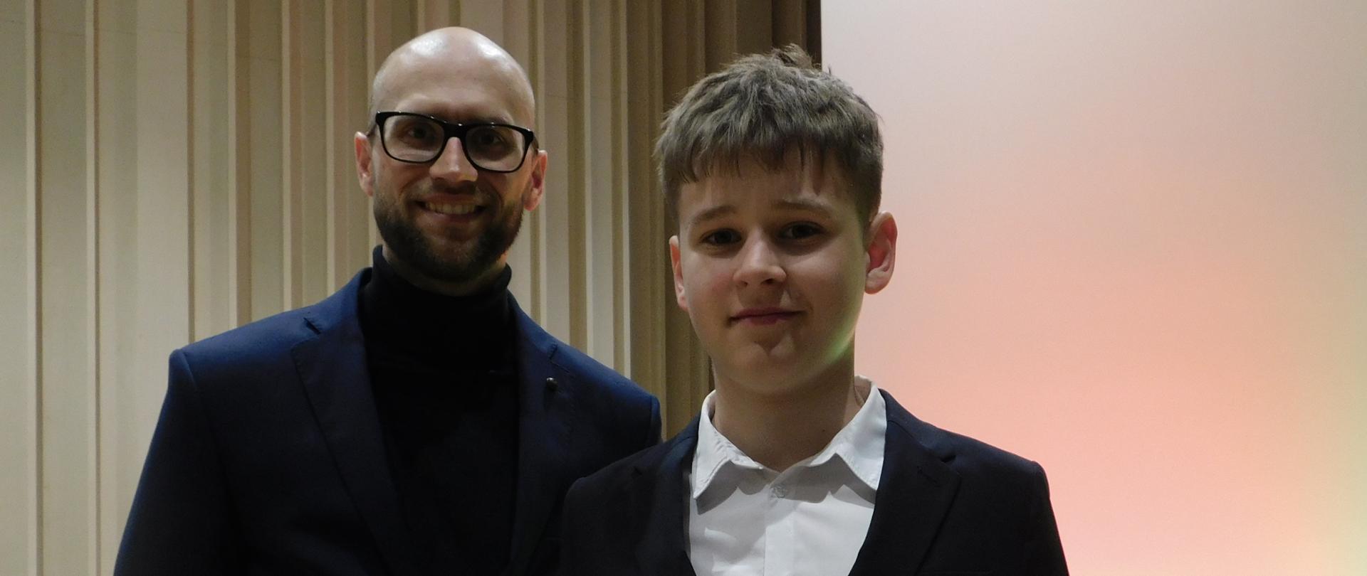 Na zdjęciu po lewej stronie łysawy mężczyzna z zarostem i w okularach. Ubrany jest w czarne spodnie i czarny golf oraz granatową marynarkę. Po prawej stronie chłopak w białej koszuli i czarnym garniturze. Chłopak Trzyma dwa dyplomy.Tło obrazka w odcieniach beżu.