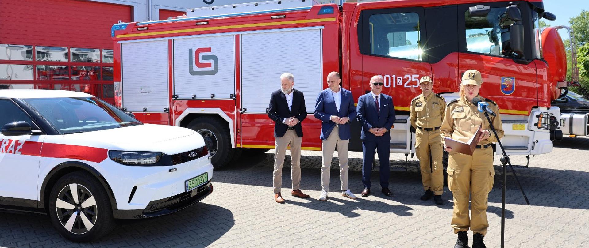 Na zdjęciu wykonanym w słoneczny dzień widać plac przed remizą strażacką. W tle znajduje się jasny budynek z dużymi czerwonymi bramami garażowymi oznaczonymi numerami „4” i „5”.
Centralną część kadru zajmuje duży czerwony wóz strażacki zaparkowany bokiem do obiektywu. Na jego boku widoczne są białe rolety skrytek na sprzęt ratowniczy. Przed wozem stoi kilka osób uczestniczących w oficjalnym wydarzeniu.
Po prawej stronie, na pierwszym planie, znajduje się umundurowana funkcjonariuszka stojąca przy mikrofonie na statywie. Trzyma w rękach dokumenty i prawdopodobnie przemawia do zgromadzonych. Za nią stoją dwaj funkcjonariusze w mundurach oraz trzej mężczyźni w garniturach lub marynarkach.
Po lewej stronie zdjęcia widoczny jest fragment białego samochodu służbowego z czerwonym pasem i napisem sugerującym przynależność do straży pożarnej.
