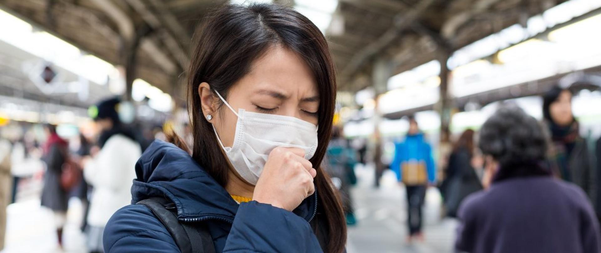 Kobieta w maseczce ochronnej stoi na peronie dworca kolejowego i kaszle w zaciśnieta dłoń. W tle widać rozmazany tłum ludzi niezwracajcych uwagi na kobiete.