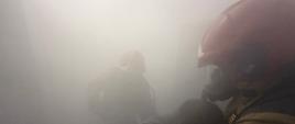 Strażak w aparacie ochrony dróg oddechowych niesie osobę poszkodowaną w silnie zadymionym pomieszczeniu; w tle zarys drugiego ratownika działającego w ograniczonej widoczności.