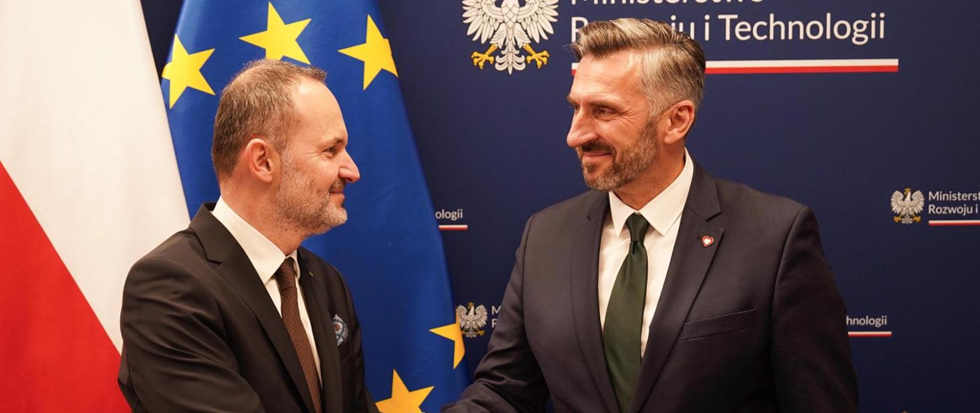 Na zdjęciu widać ministra Krzysztofa Hetmana i wiceministra Waldemara Sługockiego. W tle baner MRiT, a po prawej stronie flagi Polski i UE.