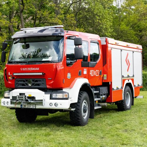 Nowoczesny, czerwony wóz strażacki Renault D15 z białym przednim zderzakiem, wyciągarką oraz zamontowaną nad grillem listwą LED (lightbar). Na boku kabiny widnieje numer „401[D]23”, a na roletach skrytek – czerwone logo Szczęśniak. Pojazd stoi na trawniku.