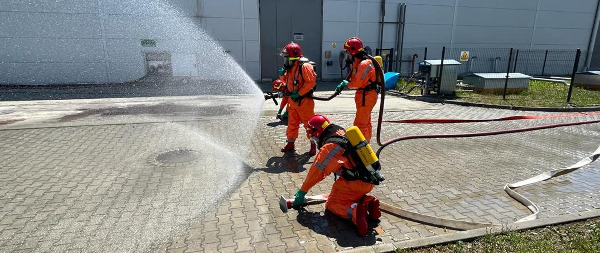 Zdjęcie ukazuje na pierwszym planie trzech strażaków, którzy są ubrani w ubrania chemiczne. Podają oni prąd wody i przygotowują kurtynę wodną. Na drugim planie widać obiekt firmy GÓRSTAL.