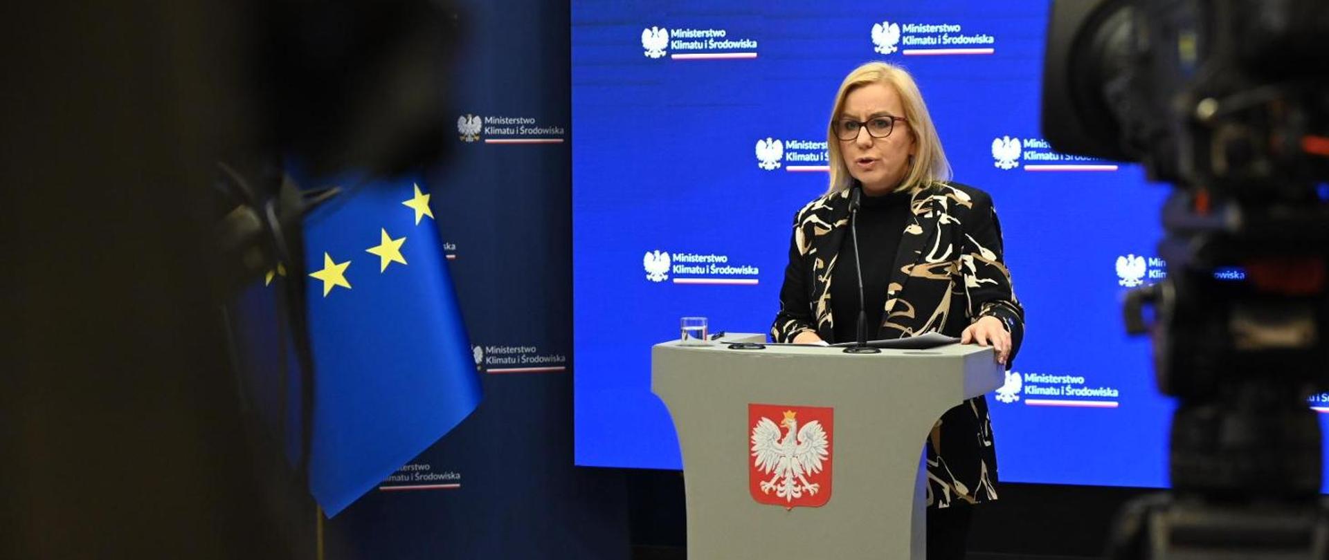 Minister of Climate and Environment Paulina Hennig-Kloska during a press briefing on irregularities in the Clean Air Programme. In the photo, the minister is standing in the center at the podium, speaking into a microphone. Cameras recording her are visible in the foreground.