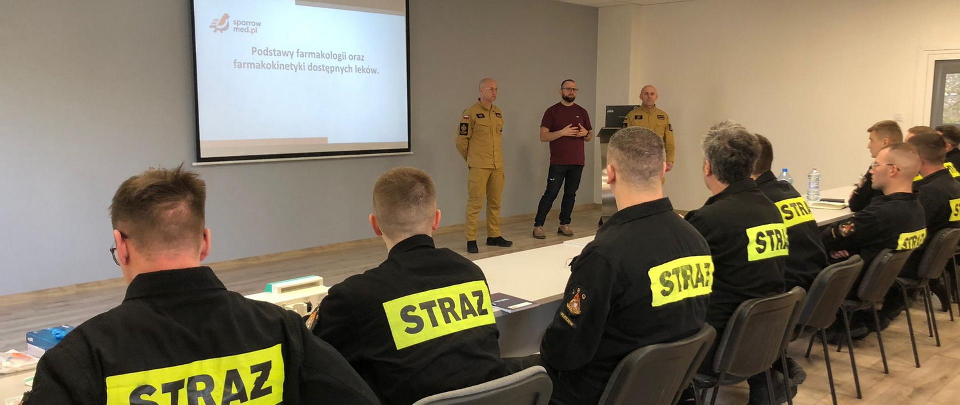 Grupa mężczyzn w mundurach straży pożarnej siedzi na oświetlonej sali, przed nimi stoi trzech mężczyzn środkowy jest ubrany cywilnie, za nimi znajduje się ekran z napisem podstawy farmakologii oraz farmakokinetyki dostępnych leków