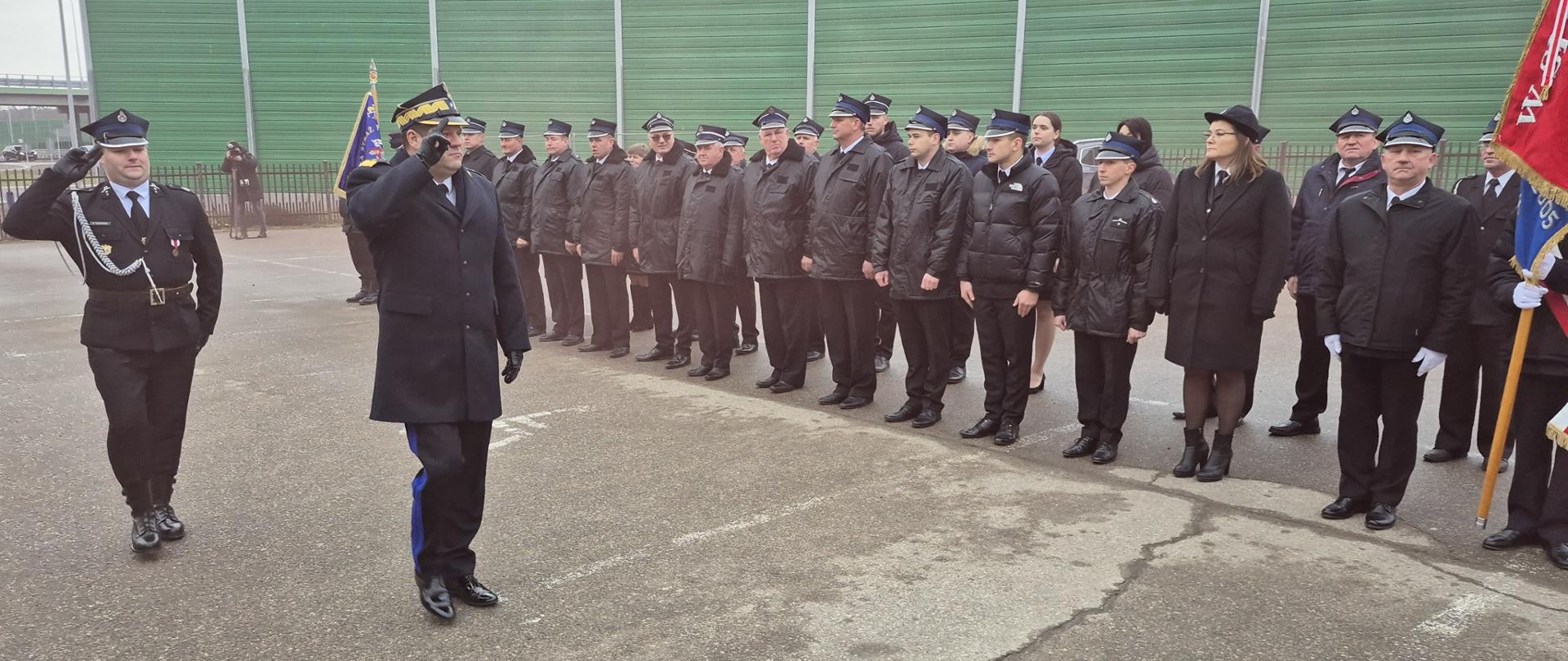 Na pierwszym planie Mazowiecki Komendant Wojewódzki PSP nadbryg. Artur Gonera w czarnym mundurze wyjściowym z płaszczem oraz strażak OSP w czarnym mundurze wyjściowym składają honor sztandarowi. Drugi plac stanowią stojący w dwuszeregu strażacy OSP w czarnych kurtkach. Stoją na placu, w tle widoczne zielone ekrany dźwiękochłonne. 