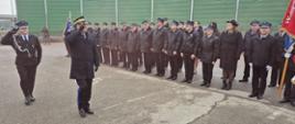 Na pierwszym planie Mazowiecki Komendant Wojewódzki PSP nadbryg. Artur Gonera w czarnym mundurze wyjściowym z płaszczem oraz strażak OSP w czarnym mundurze wyjściowym składają honor sztandarowi. Drugi plac stanowią stojący w dwuszeregu strażacy OSP w czarnych kurtkach. Stoją na placu, w tle widoczne zielone ekrany dźwiękochłonne. 