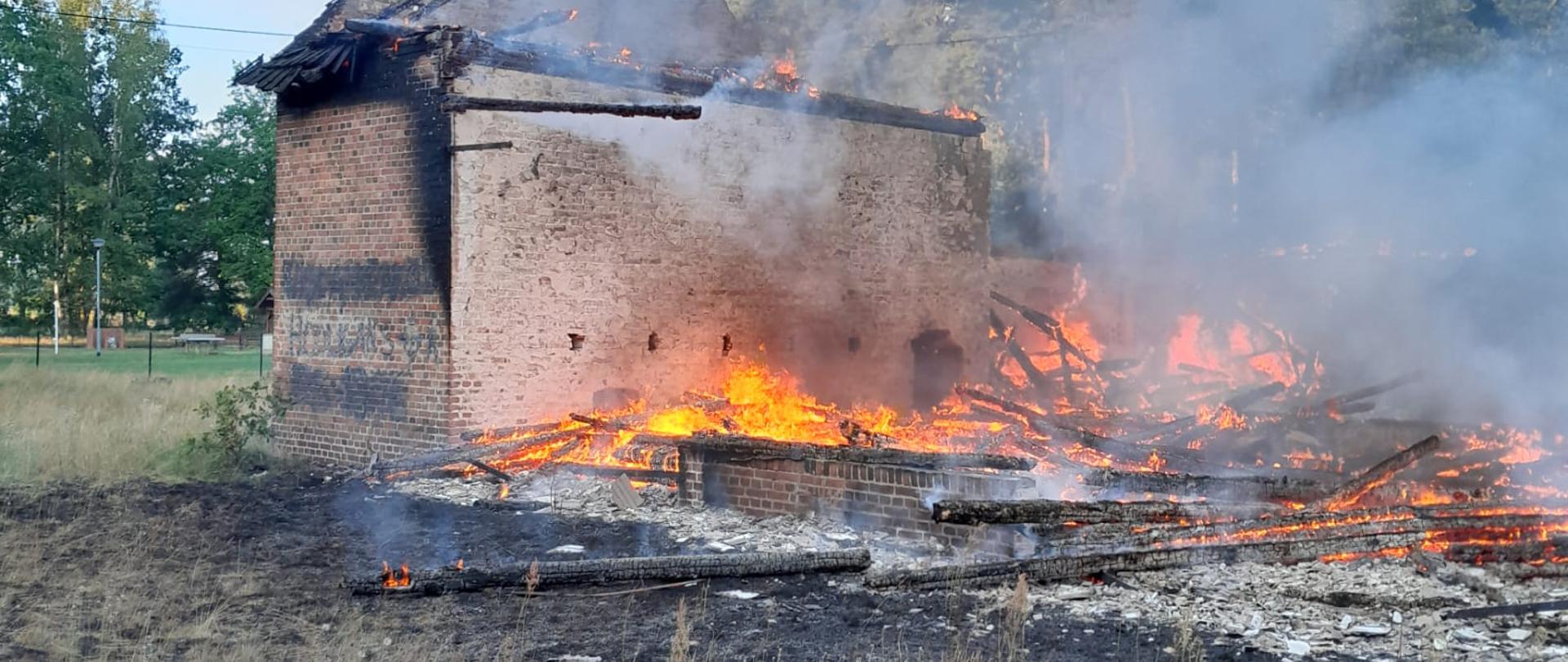 Zdjęcie przedstawia objęty pożarem budynek gospodarczy w całej objętości, który zawalił się jeszcze przed przybyciem jednostek ochrony przeciwpożarowej.