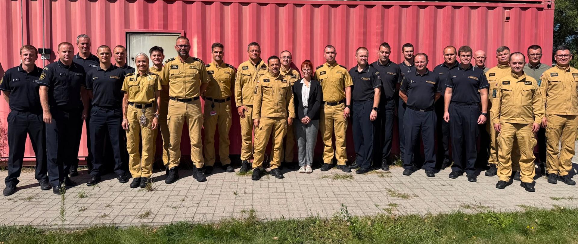 Pozowane zdjęcie grupowe uczestników spotkania na zewnątrz. Przed czerwonym kontenerem z napisem „998” stoi około trzydziestu osób – głównie strażacy w piaskowych oraz ciemnogranatowych mundurach służbowych, a także osoby w strojach cywilnych.