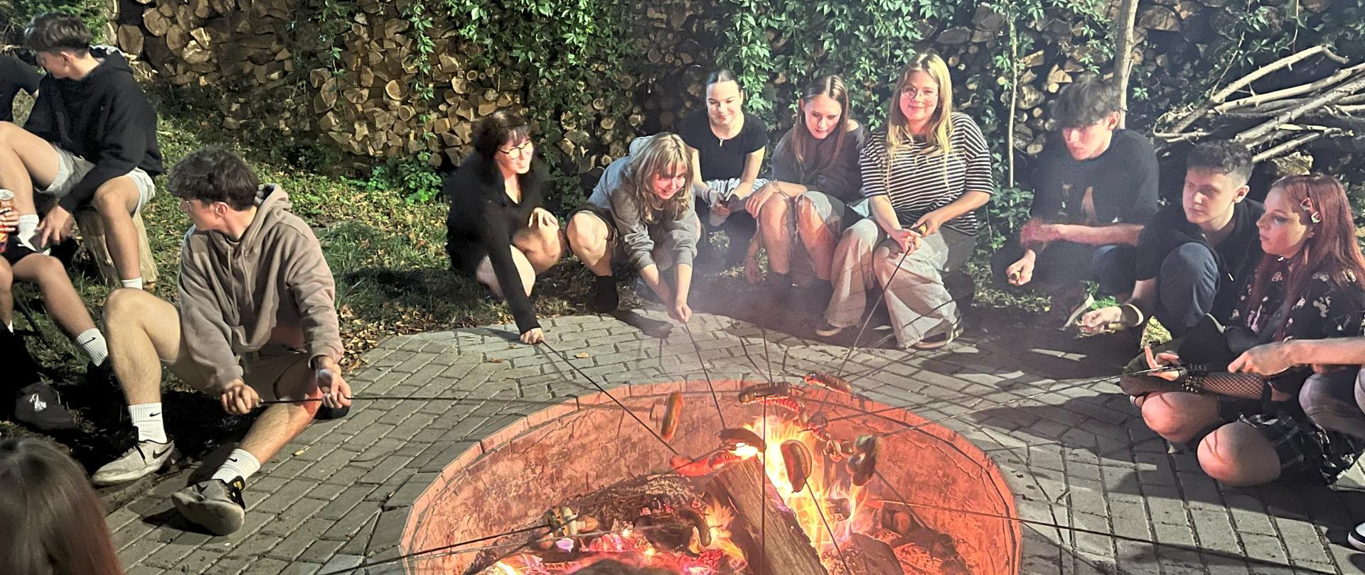 Wokół ogniska siedzi kilkanaście młodych osób, większość z nich piecze kiełbaski na kijkach. Ognisko jest otoczone szeroką opaską ułożoną z kostki brukowej. W tle widać ułożone drwa i zielone rośliny.