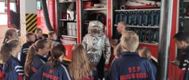 Zdjęcie przedstawia dzieci i młodzież z Młodzieżowej Drużyny Pożarniczej podczas wizyty w Jednostce Ratowniczo - Gaśnicznej. Strażak omawia przybliża pracę strażaka oraz prezentuje sprzęt strażacki. Na zdjęciu widać również samochód strażacki. 