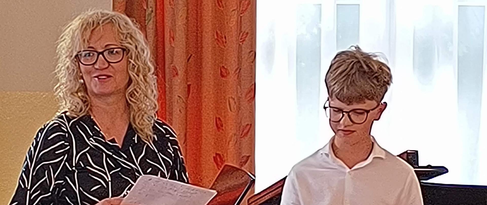 Po lewej stronie: Stoi uśmiechnięta kobieta w okularach i wzorzystej, ciemnej sukience z białymi elementami. Trzyma w rękach kartkę papieru, co sugeruje, że prowadzi wydarzenie lub wygłasza przemówienie. Po prawej stronie: Stoi młody chłopak (nastolatek) w okularach, ubrany w elegancką, białą koszulę. Wygląda na to, że właśnie wystąpił lub czeka na swoją kolej. Tuż za nim widoczny jest fragment czarnego fortepianu. Na pierwszym planie: Tyłem do obiektywu siedzi publiczność obserwująca całą sytuację. W tle widać długie zasłony oraz duże okno z jasną firanką.