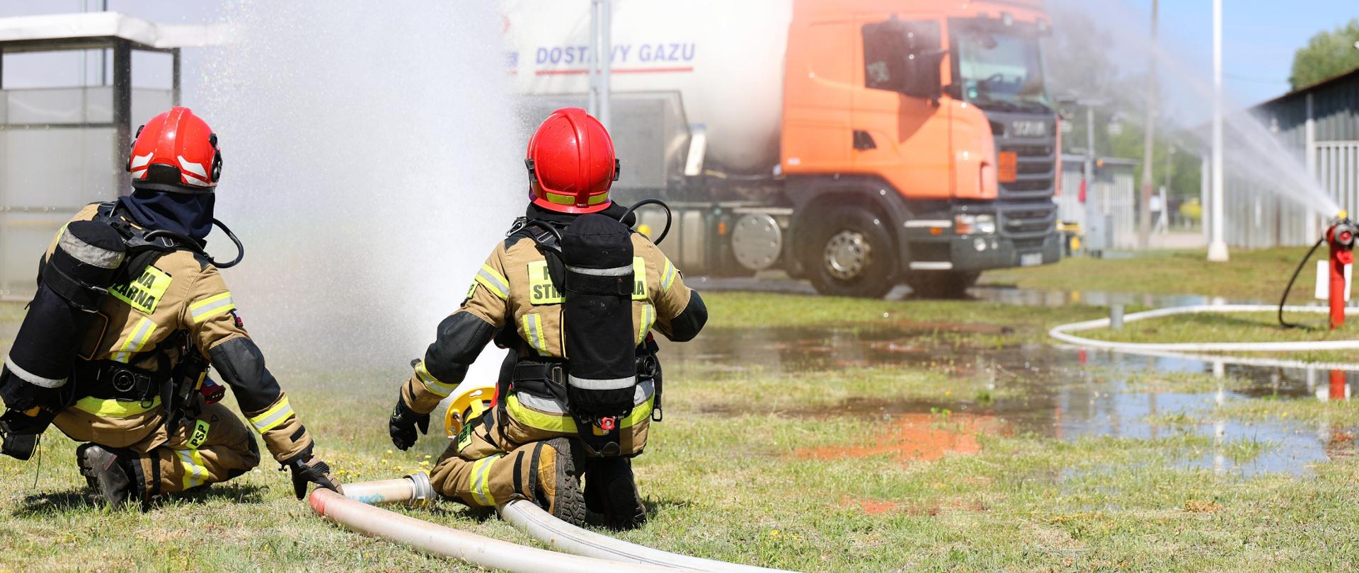 Zdjęcie przedstawia dwóch strażaków klęczących na trawie w dolnej części kadru i kierujących prąd wody w stronę instalacji przemysłowej. W centrum oraz po prawej stronie widoczna jest duża chmura rozpylonej wody. W tle stoi pomarańczowa ciężarówka oraz zbiorniki przemysłowe. Pogoda jest słoneczna i dobra widoczność sprzyja prowadzeniu ćwiczeń.