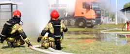 Zdjęcie przedstawia dwóch strażaków klęczących na trawie w dolnej części kadru i kierujących prąd wody w stronę instalacji przemysłowej. W centrum oraz po prawej stronie widoczna jest duża chmura rozpylonej wody. W tle stoi pomarańczowa ciężarówka oraz zbiorniki przemysłowe. Pogoda jest słoneczna i dobra widoczność sprzyja prowadzeniu ćwiczeń.