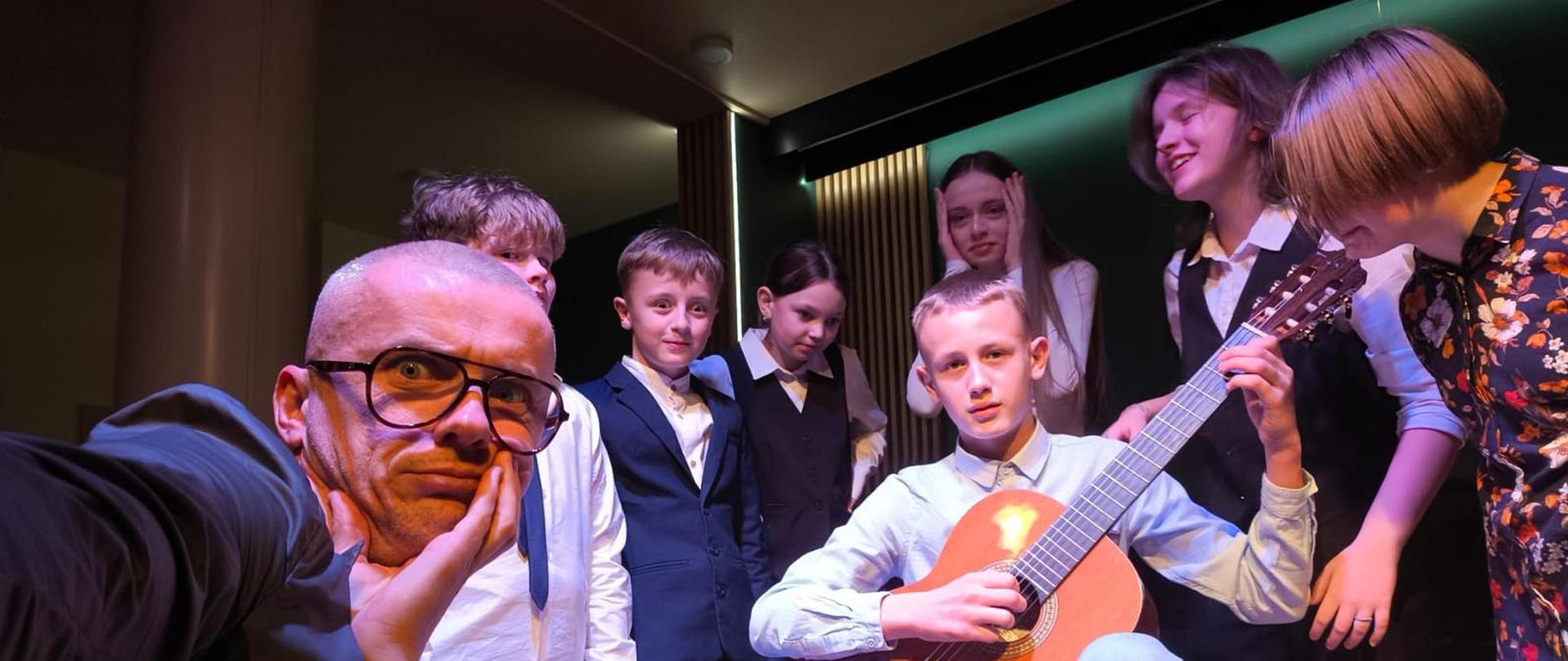 Zdjęcie przedstawia grupę młodych ludzi na scenie, otaczających chłopca grającego na klasycznej gitarze. Siedzi on na krześle, ubrany w jasnoniebieską koszulę i jeansy, a jego wyraz twarzy jest skupiony. Wokół niego stoją elegancko ubrani rówieśnicy, niektórzy się uśmiechają, inni wyrażają zdziwienie lub podziw. Na pierwszym planie dorosły mężczyzna w okularach i ciemnej marynarce robi selfie, opierając twarz na dłoni i przybierając zabawną, zaskoczoną minę. Oświetlenie sceniczne tworzy ciepłą atmosferę, a w tle widać nowoczesne wnętrze sali koncertowej z podświetlonym logo.