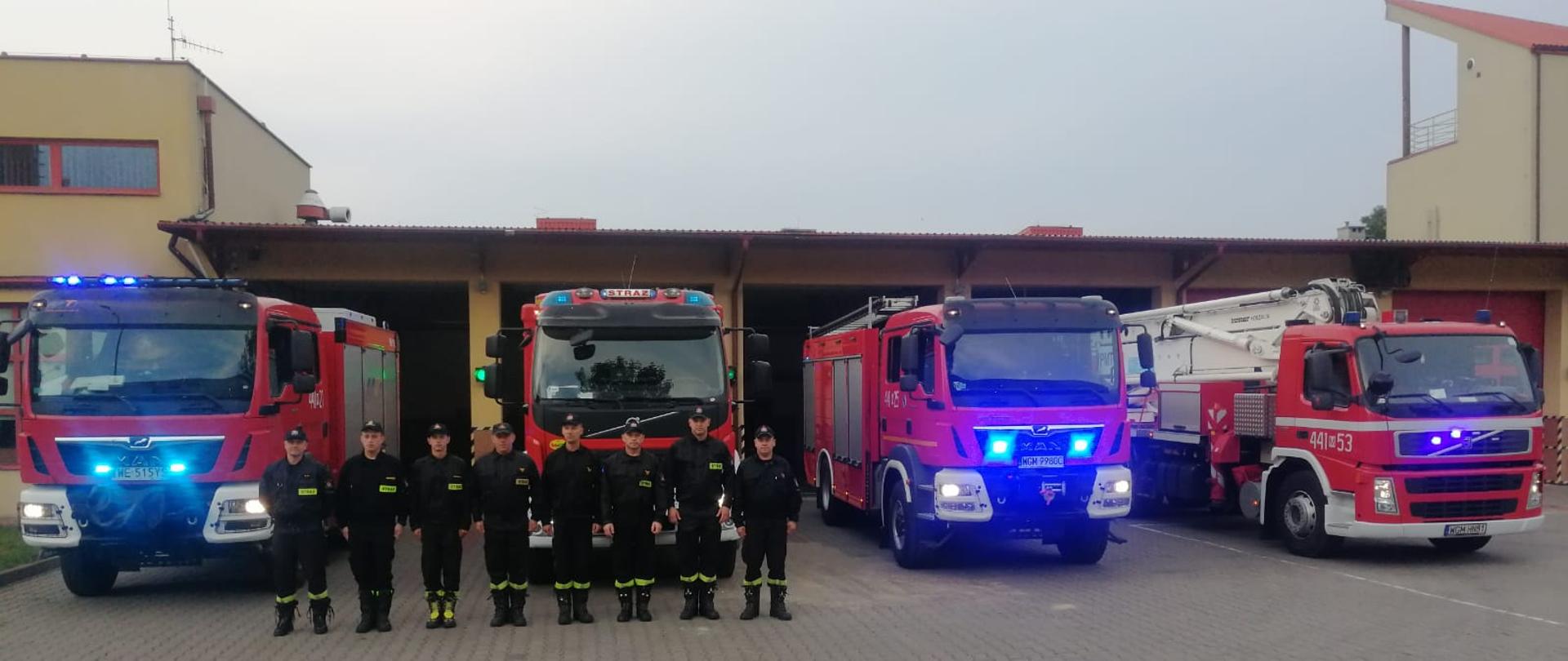 8 strażaków w ubraniach koszarowych stoi przed garażami za nimi stoją samochody ratowniczo-gaśnicze i specjalne z włączonymi sygnałami świetlnymi i dźwiękowymi w celu uczczenia pamięci tragicznie zmarłego strażaka OSP.