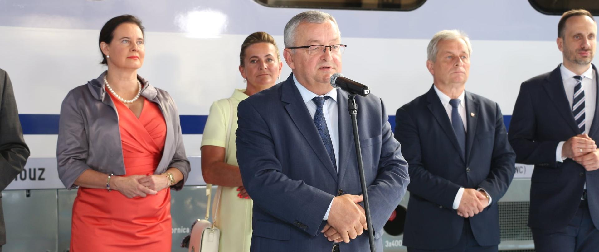 Na zdjęciu przemawiający na stojąco minister infrastruktury, który ubrany jest w stalowy garnitur - przemawia za mikrofonem na statywie. Mężczyzna ma siwe włosy, a jego oczy zdobią okulary w metalowych ramkach. W tle uczestnicy konferencji, tj. dwie panie poseł stojące po lewej stronie kadry, wojewoda i jeden z posłów. W dalszej perspektywie wagon pociągu. 