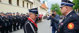 Widok z boku. Członek Komisji Rewizyjnej ZG ZOSP RP wręcza złoty znak związku zasłużonemu druhowi z OSP Kaczyna. W tle pododdział OSP. 
