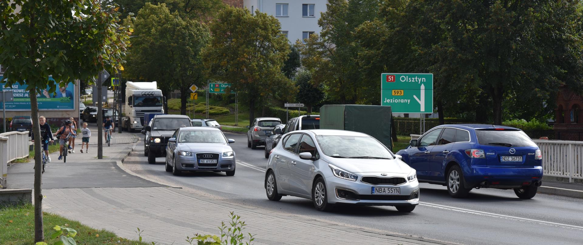 Na zdjęciu widoczna jest ruchliwa ulica, po której porusza się kilka pojazdów, w tym biały samochód na pierwszym planie, niebieski samochód oraz ciężarówka. Po prawej stronie znajduje się zielony znak drogowy wskazujący kierunki do Olsztyna i Jezioran drogą krajową nr 51. W tle widać drzewa, zabudowania oraz wieżę z zegarem. Po lewej stronie ulicy spacerują piesi i poruszają się rowerzyści.