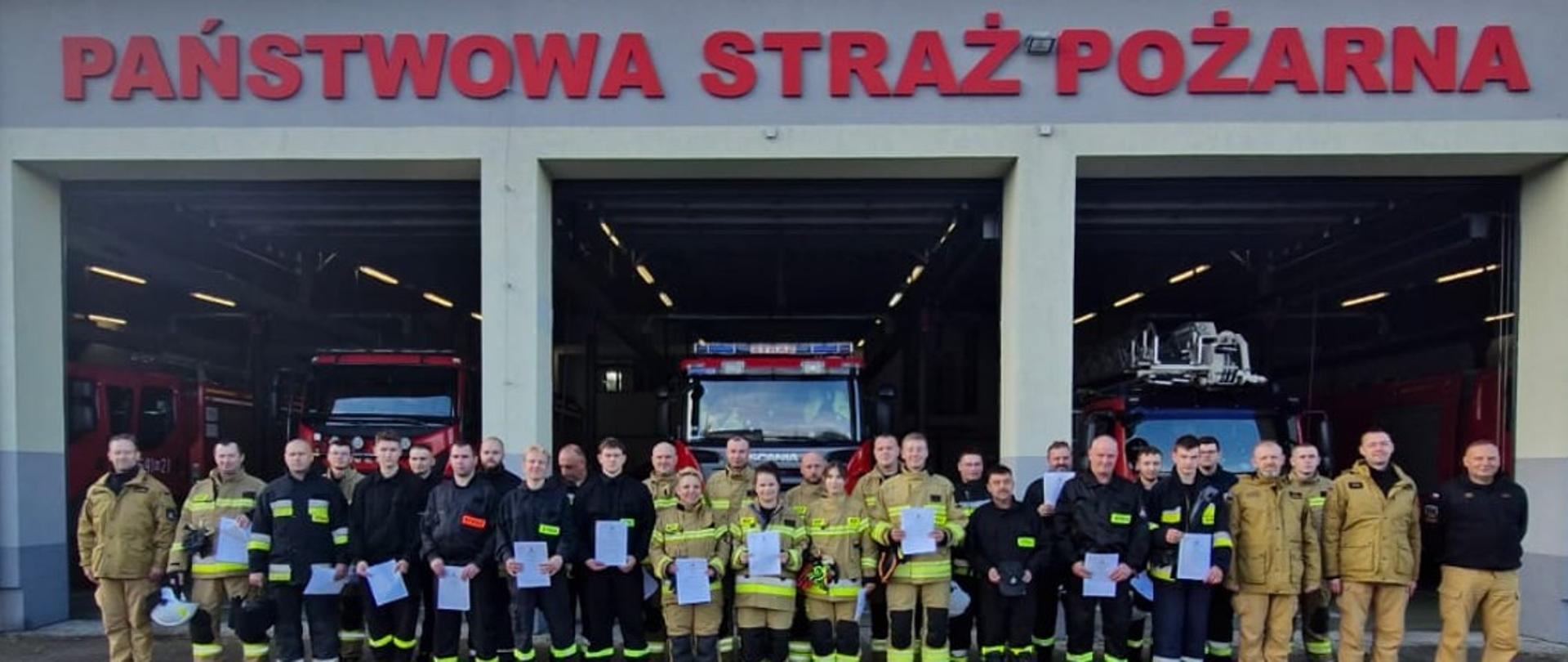Zdjęcie przedstawia grupę strażaków, stojących na zewnątrz przed budynkiem straży pożarnej.
Lokalizacja: W tle widoczny jest budynek z napisem "PAŃSTWOWA STRAŻ POŻARNA". Poniżej znajdują się otwarte bramy wjazdowe, za którymi stoją wozy strażackie (jeden z nich, centralnie umieszczony, jest wyraźnie widoczny).
Osoby: W pierwszym rzędzie stoi duża grupa (około 30) dorosłych osób. Większość z nich ubrana jest w mundury strażackie, w tym kombinezony i kurtki w kolorach beżowo-żółtym i ciemnym/czarnym.
Detale: Wiele osób trzyma w rękach certyfikaty ukończenia szkolenia podstawowego OSP. Wśród widocznych osób są zarówno kobiety, jak i mężczyźni.
Nastrój: Uczestnicy stoją z dumnymi i poważnymi minami, tworząc formalne zdjęcie grupowe.