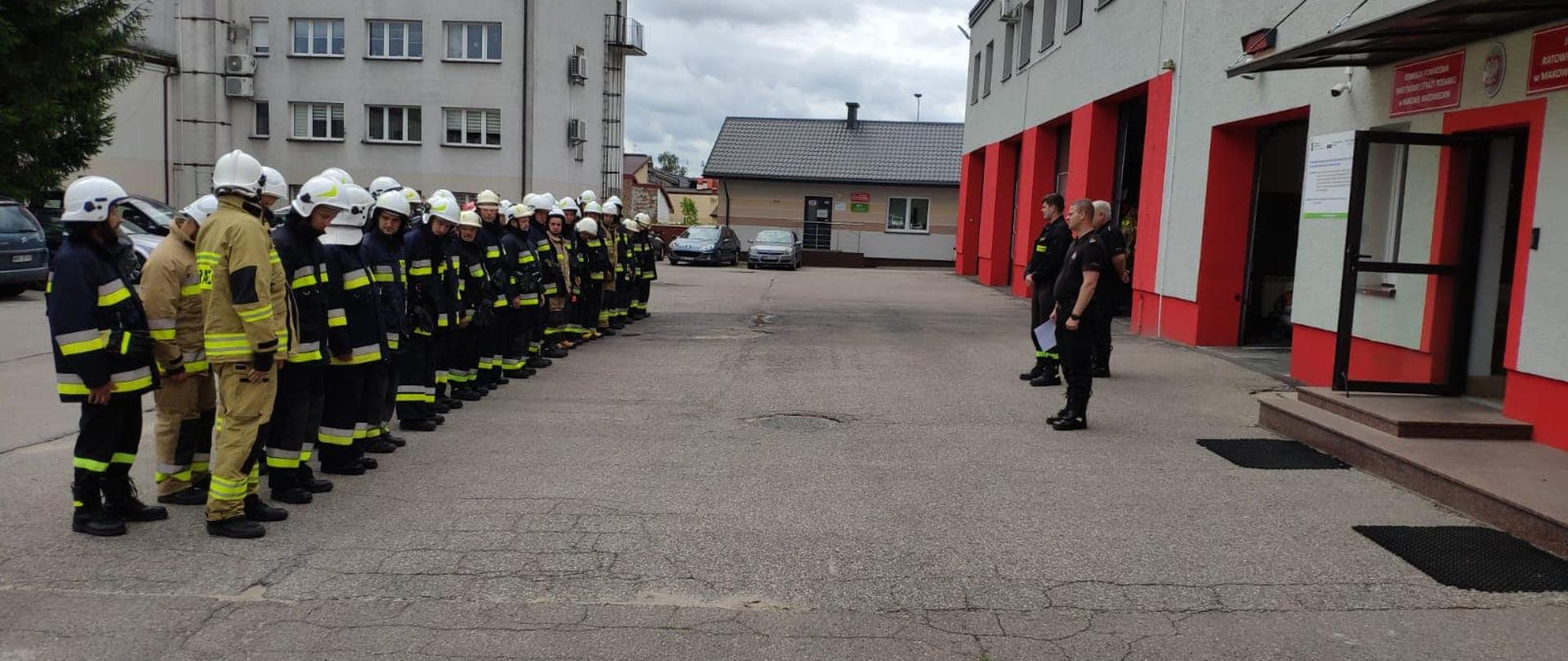 Strażacy OSP stoją w dwuszeregu na placu Komendy frontem do budynku strażnicy. Przed nimi stoi 3 funkcjonariuszy PSP.