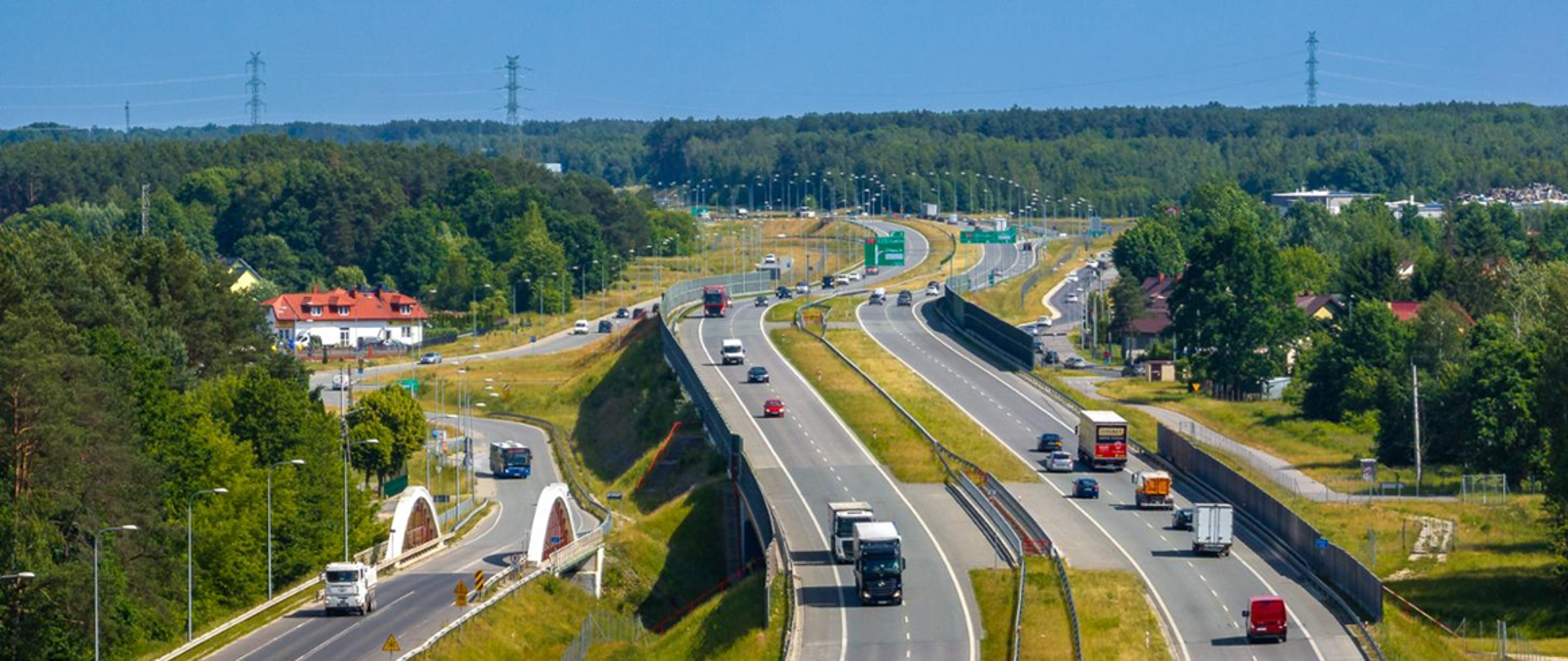 Widok z lotu ptaka na drogę ekspresową z dwoma jezdniami i ruchem pojazdów, przebiegającą przez tereny leśne i zabudowę, widoczne węzły drogowe, ekrany akustyczne oraz drogi dojazdowe