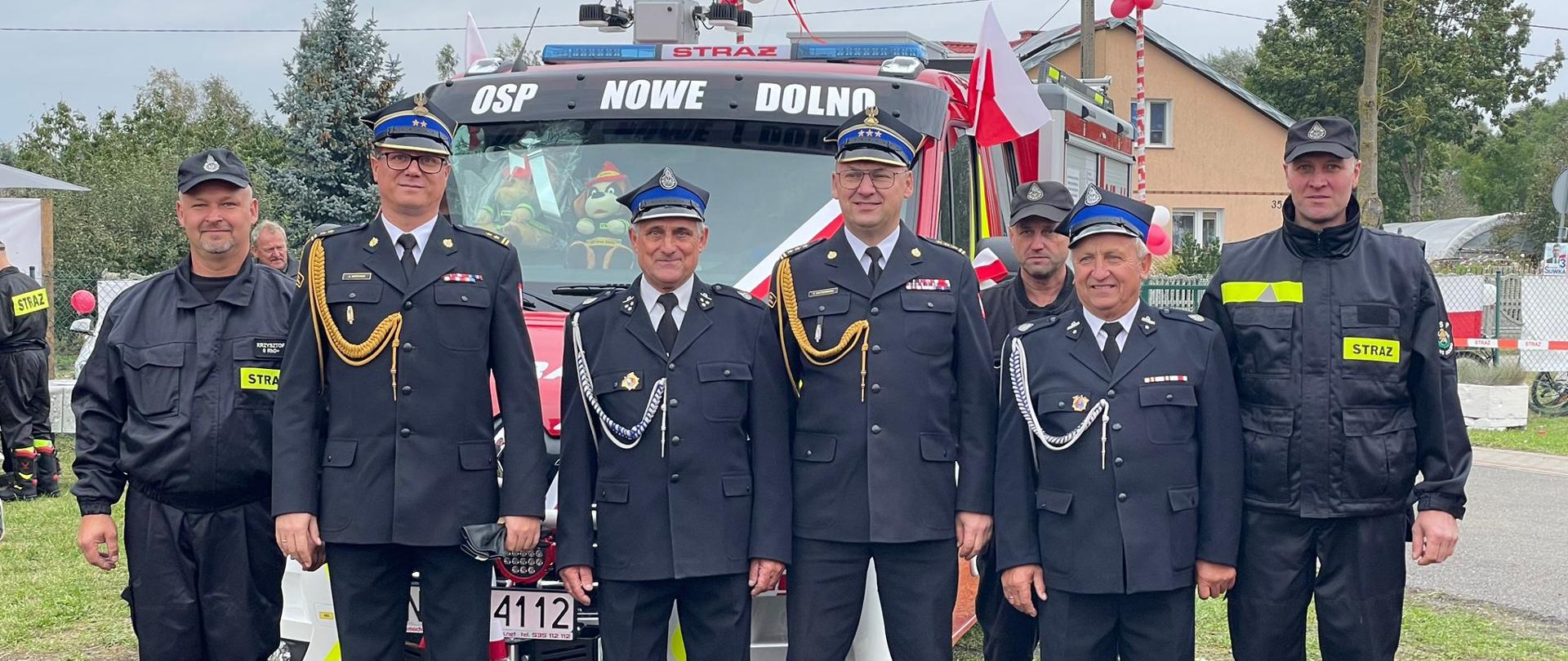 Strażacy ochotnicy z Nowego Dolna stoją w towarzystwie komendanta miejskiego państwowej straży pożarnej z Elbląga na tle nowego pojazdu pożarniczego. W tle budynki mieszkalne.