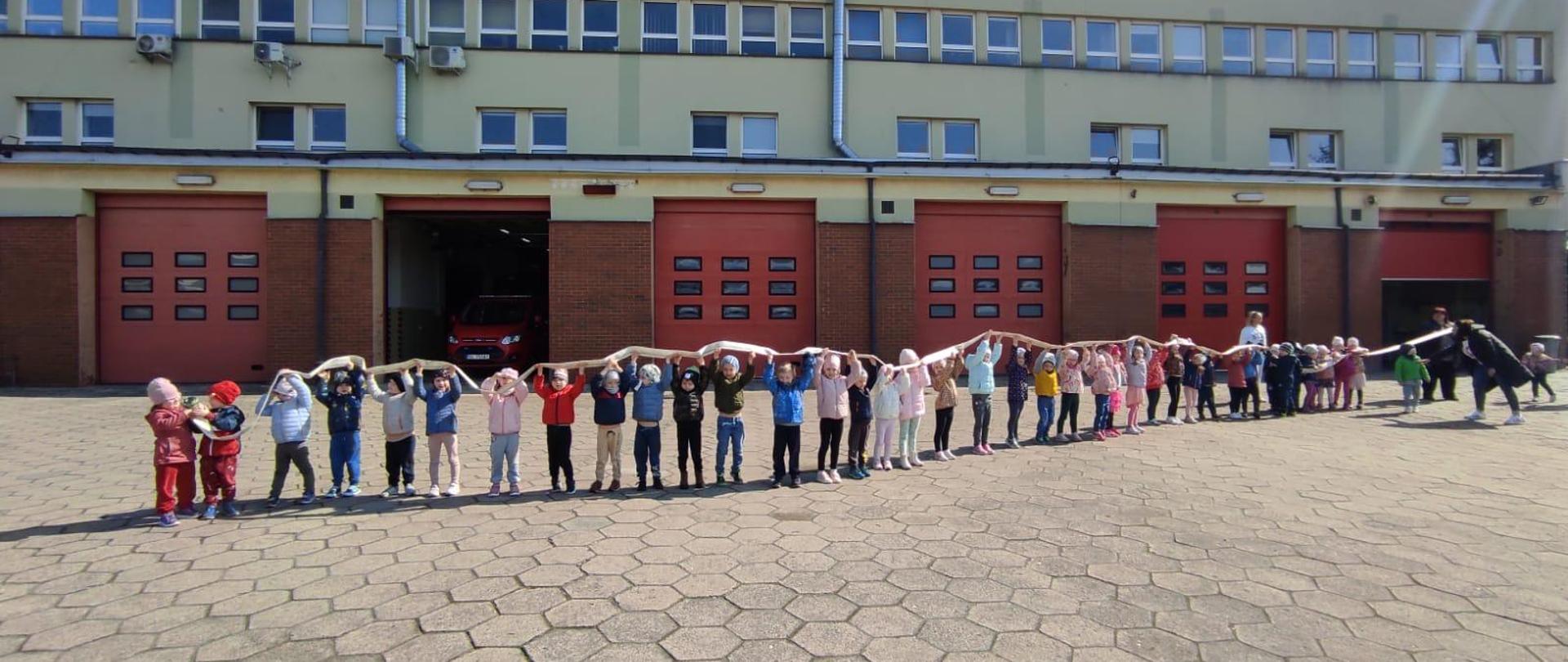 Grupa dzieci trzymająca w górze wąż pożarniczy. Stojąca na placu przed budynkiem straży pożarnej
