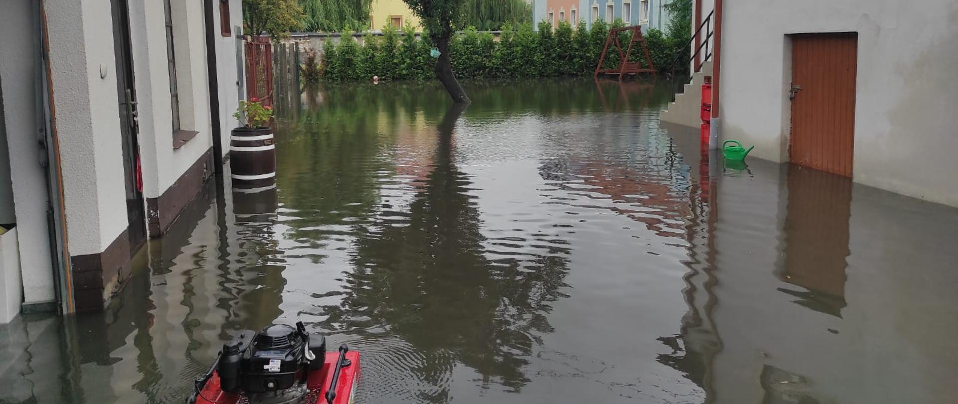 Widok zalanego podwórza. Po prawej i lewej stronie budynki Na wodzie pływająca pompa strażacka z podłączonym wężem.