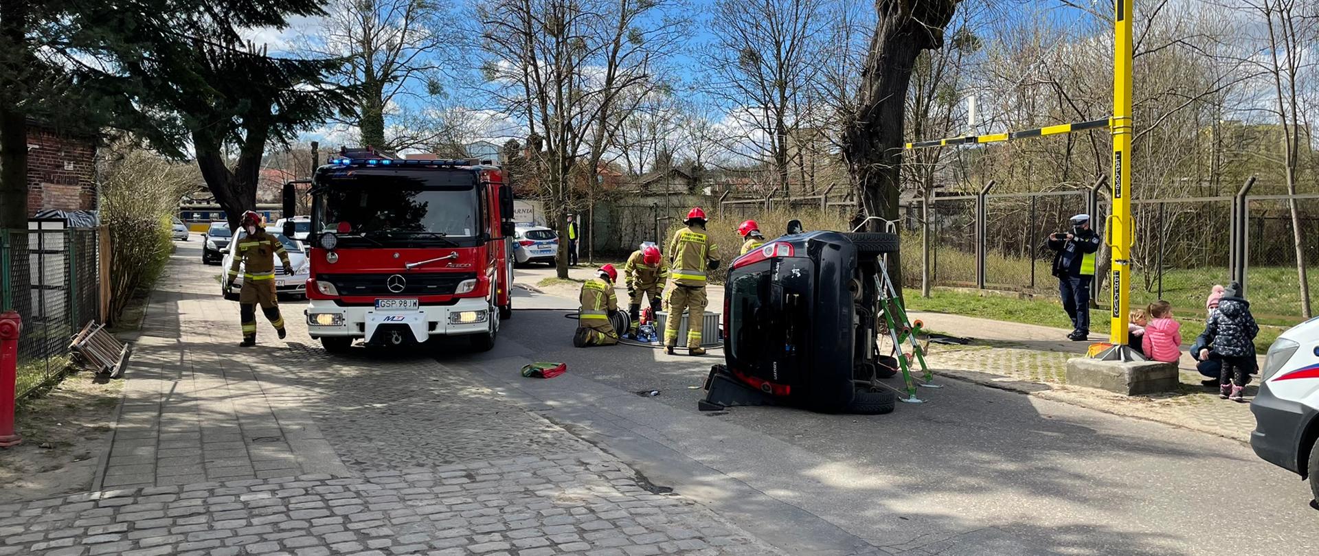 Samochód osobowy po wypadku leży na boku. Na zdjęciu widać Policjanta wykonującego zdjęcia miejsca zdarzenia do dalszych czynności.
