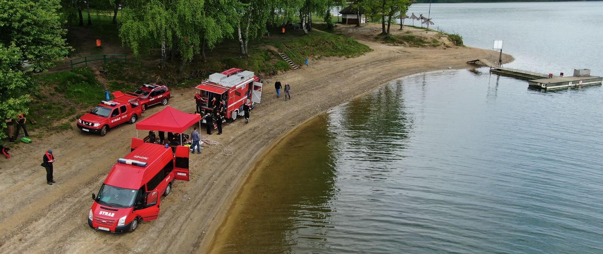 Na zdjęciu strażacy nurkowie oraz specjalne samochody strażackie na brzegu jeziora solińskiego, między samochodami stanowisko do pracy operatora systemu przewodowego pod rozłożonym namiotem, zdjęcie wykonane z drona