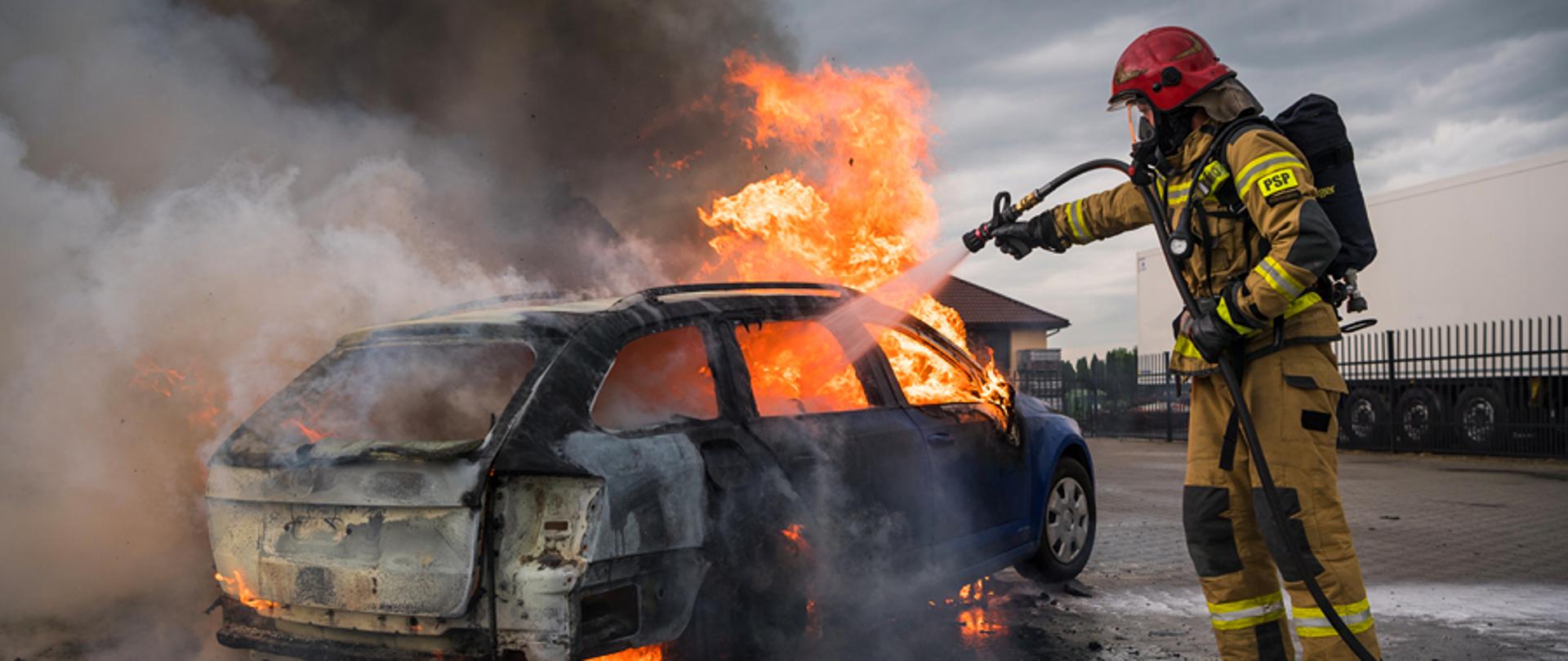 Na zdjęciu widoczny samochód osobowy koloru niebieskiego w nadwoziu typu kombi w całości objęty pożarem. Po prawej stronie kadru strażak w ubraniu specjalnym koloru piaskowego, czerwonym hełmie, masce na twarzy i aparatem powietrznym na plecach, gasi pożar prądem wody.