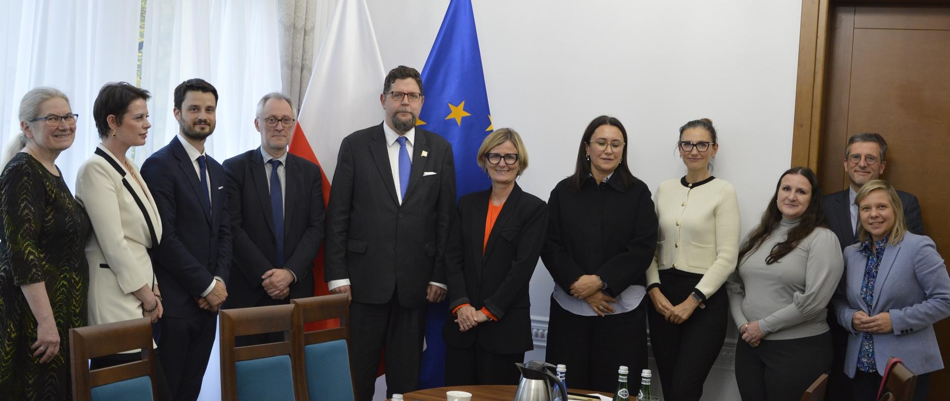 Pod ścianą stoi 11 osób, pośrodku grupy wiceminister Szeptycki, za nimi pod ścianą flagi Polski i UE, z boku drewniane drzwi.