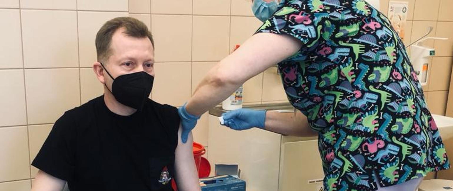 Zdjęcie przedstawia funkcjonariusza Państwowej Straży Pożarnej w pomieszczeniu szpitalnym punktu szczepień podczas wykonywanej iniekcji dawki szczepionki. Funkcjonariusz siedzi na krześle szpitalnym ubrany w mundur koszarowy oraz maseczkę jednorazową. Pielęgniarka stojąca z boku dokonuje szczepienia.