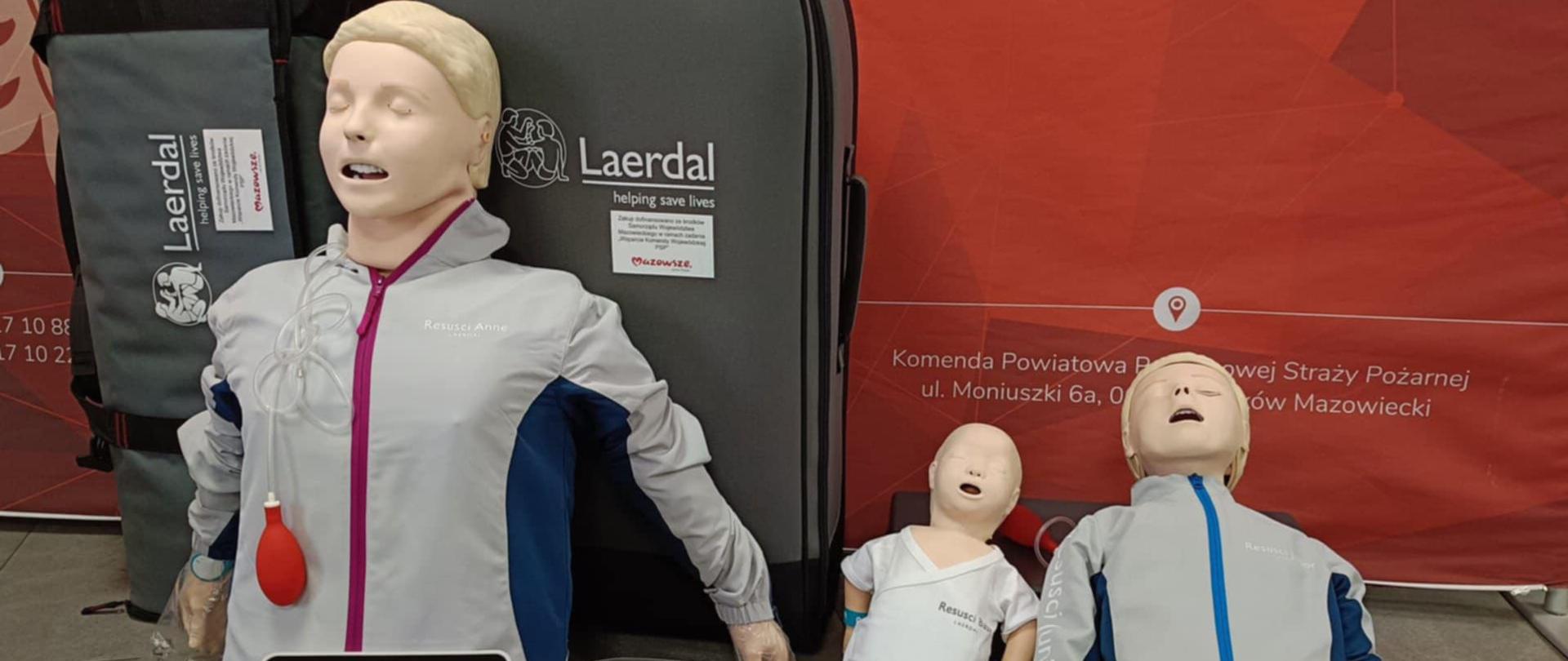 Zdjęcie przedstawia trzy fantomy ratownicze (osoba dorosła, dziecko, niemowlak) ubrane w bluzy i spodnie. Na zdjęciu znajdują się także pokrowce od fantomów