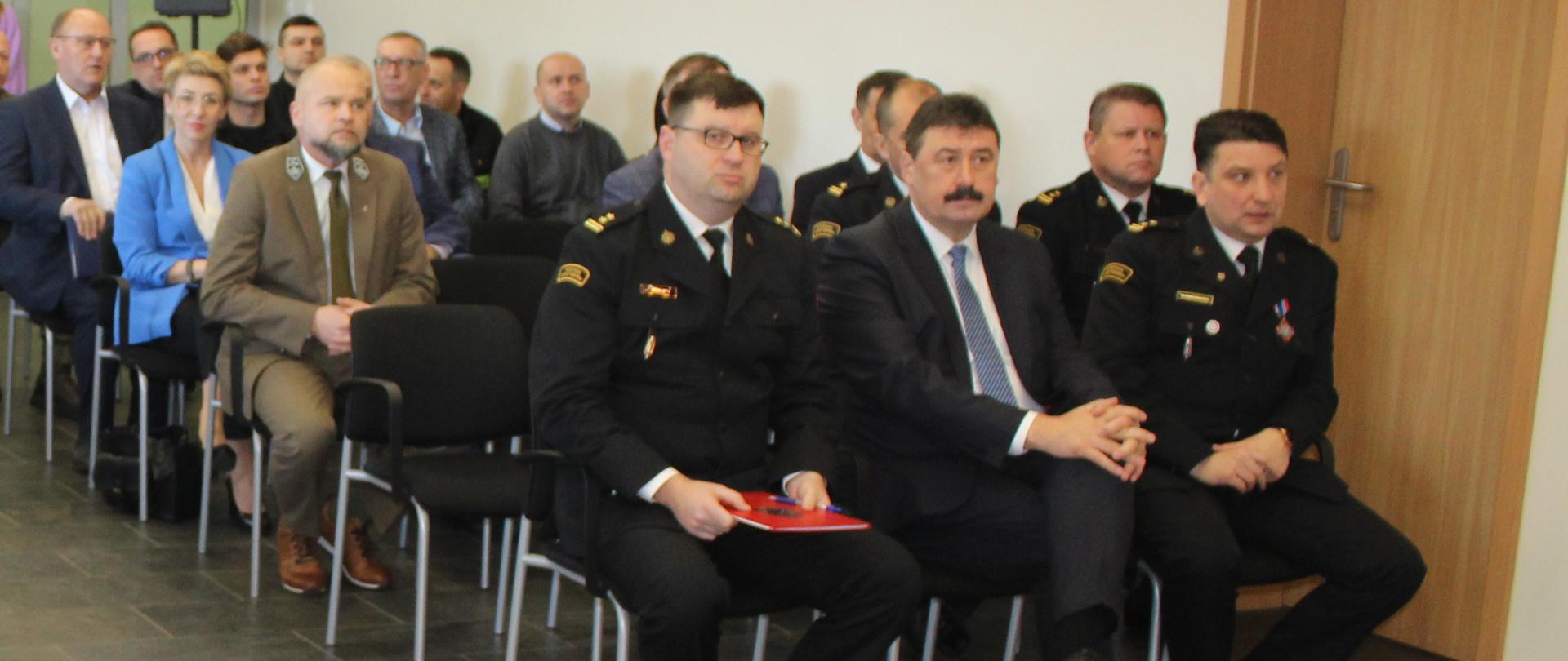 grupa ludzi siedzi w pomieszczeniu w dwóch rzędach, z przodu siedzą strażacy