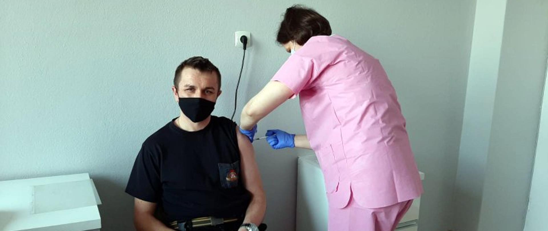 Na zdjęciu widać siedzącego strażaka w ciemnym mundurze koszarowym, który właśnie przyjmuje szczepionkę przeciw COVID-19. Po prawej stronie stoi pielęgniarka, która wykonuje zastrzyk.