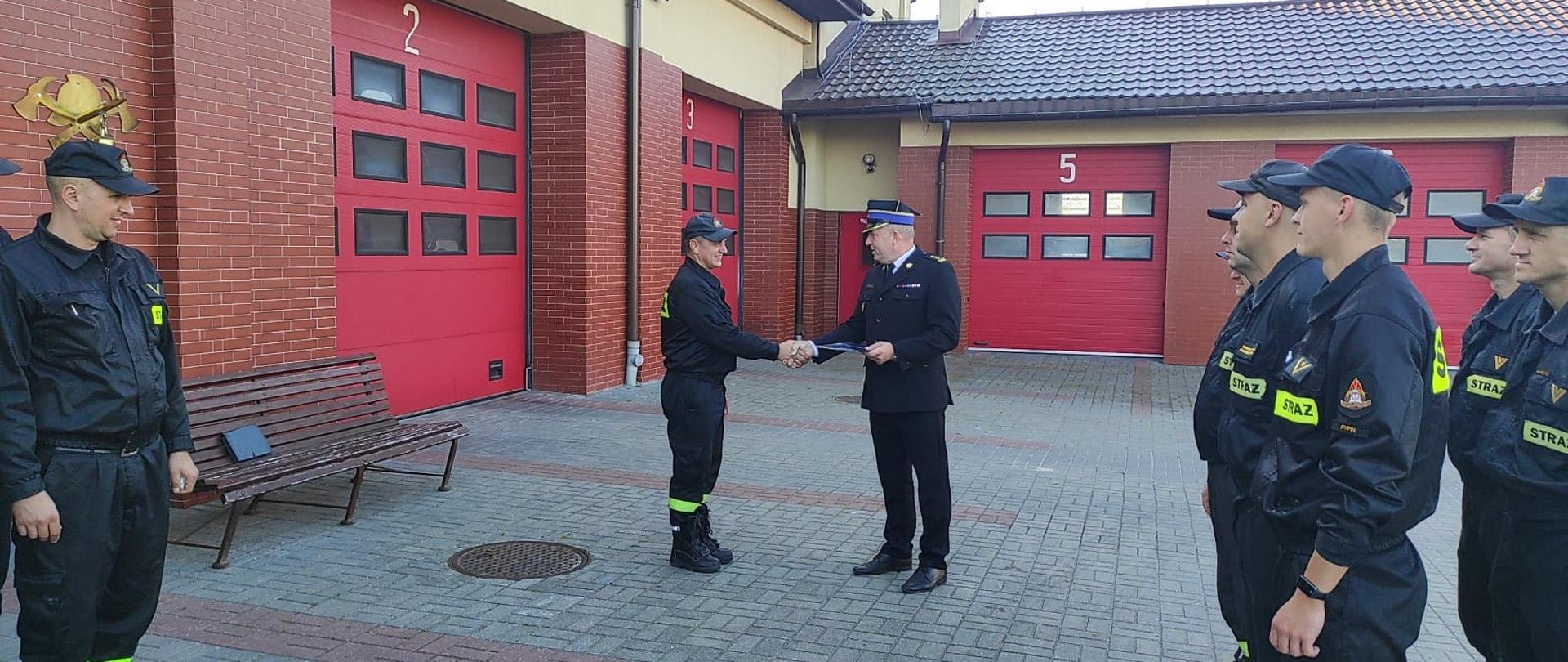 Komendant Powiatowy PSP w Rypinie starszy brygadier Grzegorz Łydkowski wręcza list gratulacyjny z okazji jubileuszu służby starszemu ogniomistrzowi Andrzejowi Szrull. Stoją oni na wprost siebie. Za nimi czerwone wrota garażowe. Po prawej i lewej stronie na polbruku stoją strażacy w dwuszeregach.