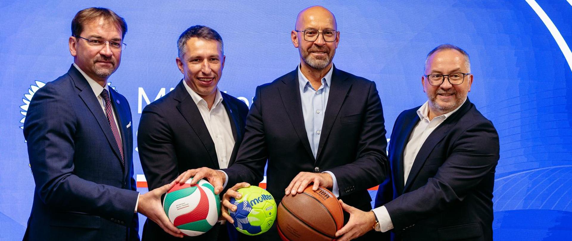 Prezes Polskiego Związku Piłki Siatkowej Sebastian Świderski, prezes Związku Piłki Ręcznej w Polsce Sławomir Szmal, minister sportu i turystyki Jakub Rutnicki i prezes Polskiego Związku Koszykówki Grzegorz Bachański stoją na błękitnym tle, trzymając piłki do siatkówki, piłki ręcznej i koszykówki.
