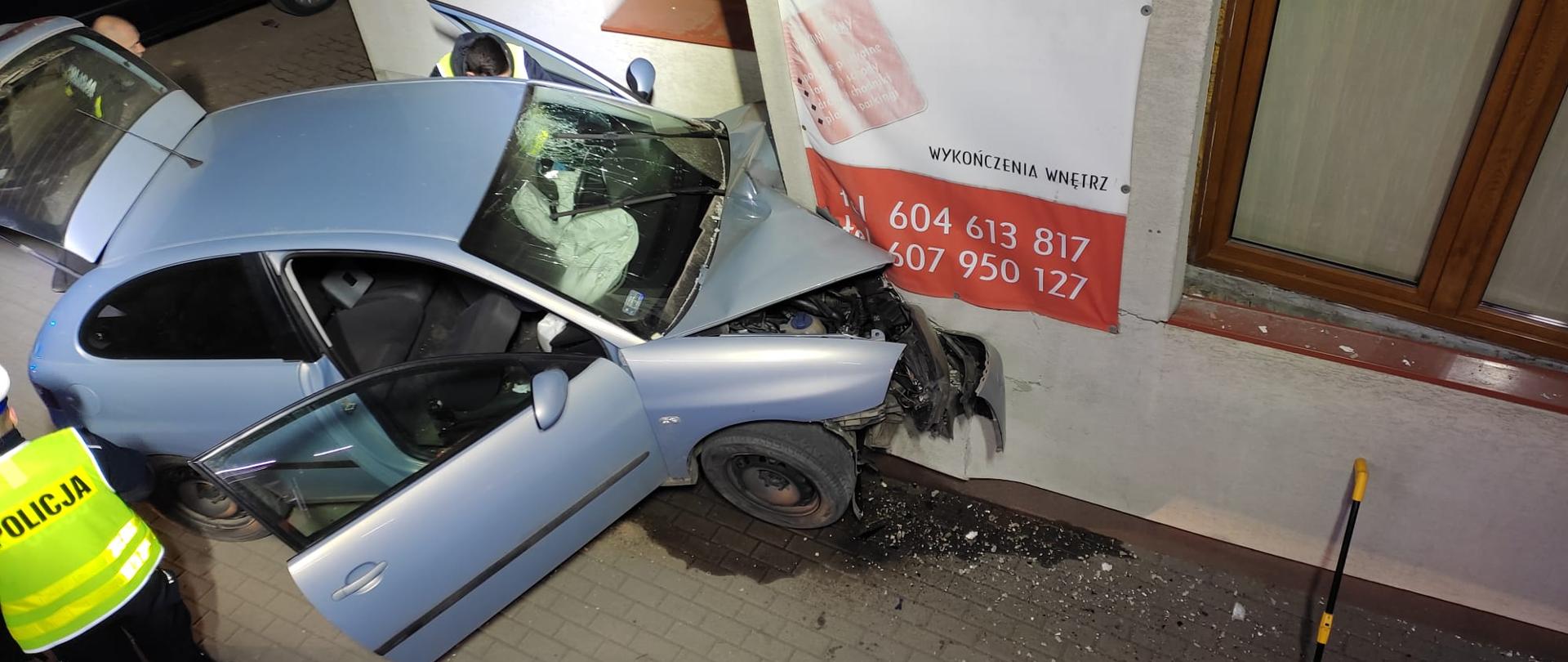 Zdjęcie przedstawia samochód osobowy który uderzył w ścianę budynku. Samochód ma otwarte przednie prawe i lewe drzwi oraz tylna klapę. Przód pojazdu jest wbity w ścianę budynku mieszkalnego. po lewej stronie tyłem stoi Policjant w seledynowej kamizelce. Za otwarta klapa tylną widać drugiego Policjanta a przy lewych otwartych drzwiach jest trzeci policjant. Po prawej stronie stoi przy ścianie urządzenie do mierzenia odległości. Po lewej stronie widać czarny samochód osobowy. Na ścianie budynku wisi baner reklamowy z napisem brukarstwo